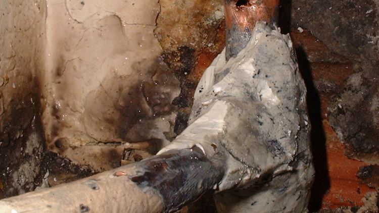 A corroded pipe joint wrapped in a hardened patch against a stained, damp wall