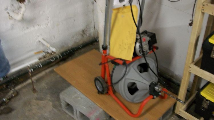 An electric drain cleaning machine with its cable drum, standing on a board in a basement