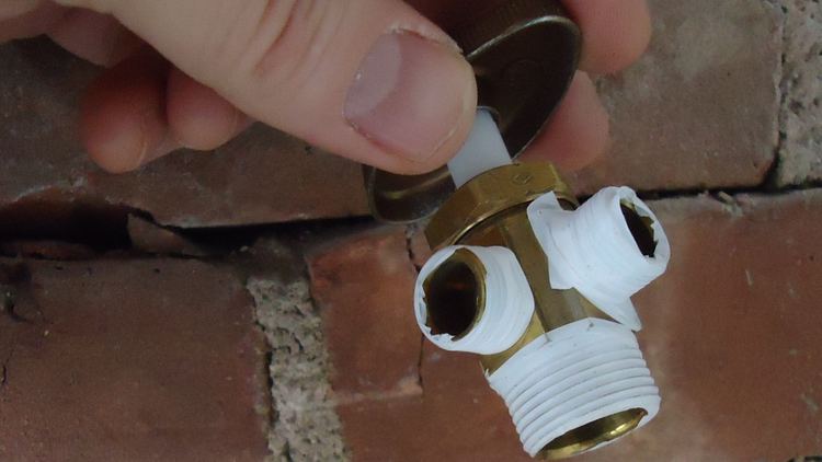 A hand holding a brass stop valve with white thread tape against a brick wall